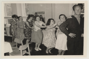 Eine Gruppe von Menschen, die in einem Raum mit Stühlen und einem Tisch tanzen, der mit einer weißen Decke bedeckt ist, mit einer Wand, die ein Photo und Fenster im Hintergrund zeigt, und den Anschein erweckt, aus den 1950er Jahren zu stammen.