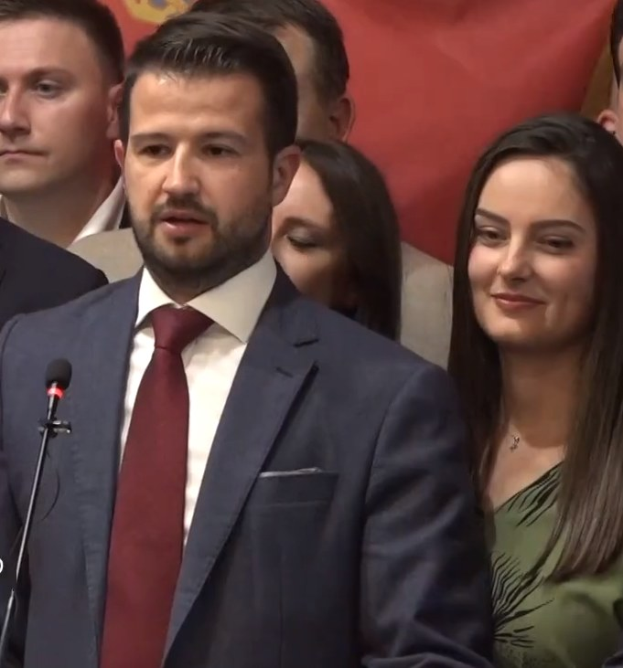 Ein Mann in Anzug und Krawatte spricht in ein Mikrofon an einem Podium, mit einem Banner dahinter und einer Menge davor, wahrscheinlich zu einem Verbot der sozialen Medien durch die Regierung.