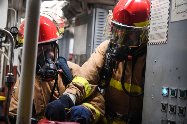 Zwei Feuerwehrleute in Schutzausrüstung arbeiten an einem Feuerwehrauto, wobei eine Tafel und Metallstangen im Hintergrund sichtbar sind.