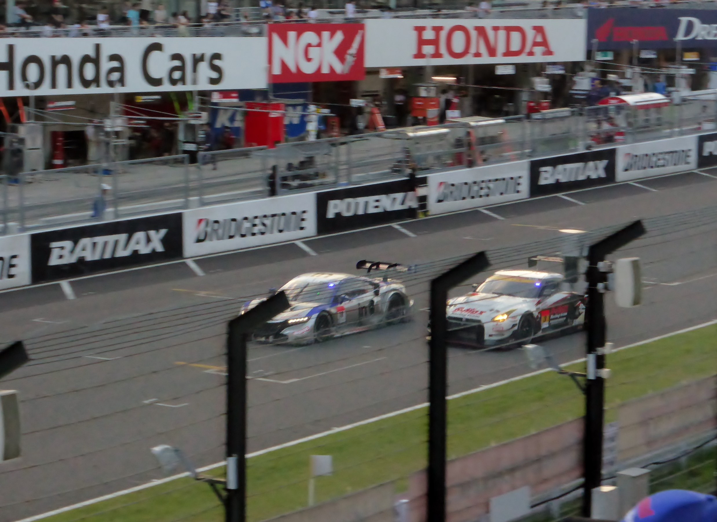 Eine Gruppe von Autos, die auf einer Rennstrecke mit Absperrungen, Beschilderung, Barrieren und umgebenen Strukturen fahren.