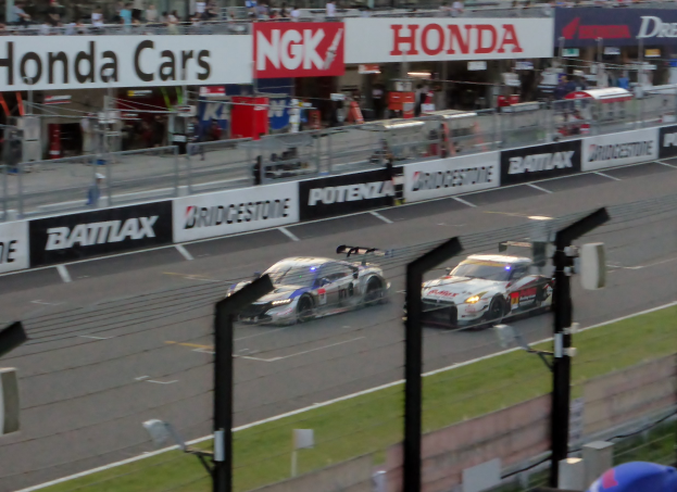 Eine Gruppe von Autos, die auf einer Rennstrecke mit Absperrungen, Beschilderung, Barrieren und umgebenen Strukturen fahren.