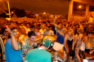 Menschen tanzen vorne bei einem Konzert mit Lichtern, Pfählen, Bäumen und anderen Objekten im Hintergrund.