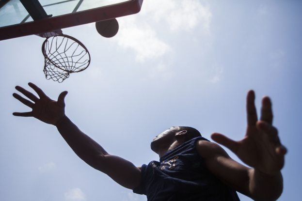 Ein Mann in einem Basketballtrikot und Shorts springt, um einen Basketball in einen Korb unter einem klaren blauen Himmel zu werfen, Arme ausgestreckt.