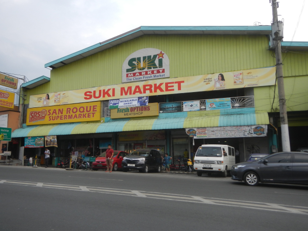 Eine belebteStraßenszene inSan Roque,Philippinen,mitFahrzeugen,Passanten,GebäudenmitSchildern,StrommastenundewolkigemHimmel,mitdemSukiMarketinderMitte.