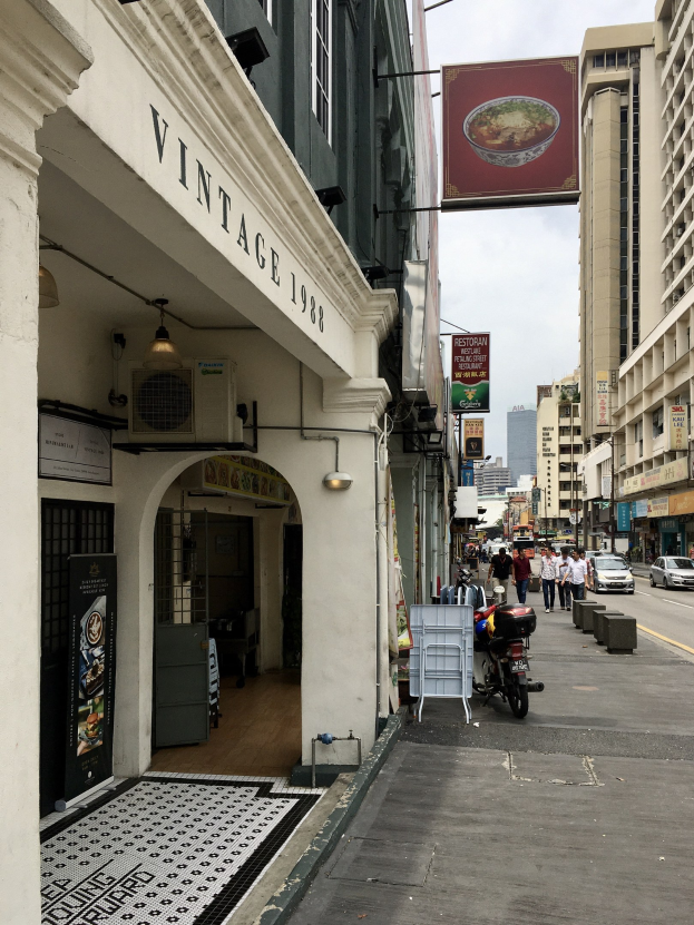 Eine belebte Straßenszene in Singapur mit Fahrzeugen, Fußgängern und Gebäuden unter einem bewölkten Himmel, mit einem Gebäude mit einer Beschriftung auf der linken Seite.