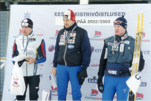 Drei Männer in Wintersporterkleidung stehen auf einem Podest, jeder hält ein Paar Skier und ein Blatt Papier in der Hand, mit einem Banner im Hintergrund.
