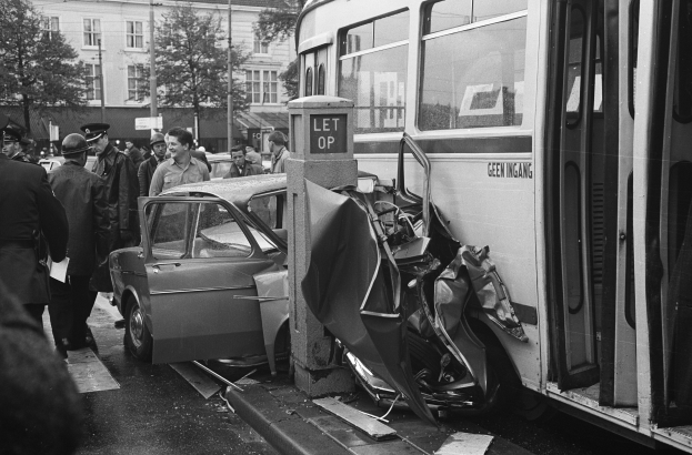 Black and white image of a bus that has crashed into a car on the side of the road with people standing around the scene and trees and buildings in the background.