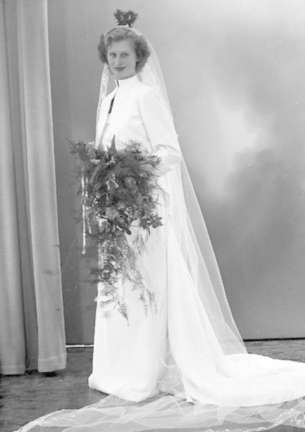 Eine Frau in einem weißen Hochzeitskleid mit Schleier und Hochsteckfrisur steht mit einem Strauß in der Hand da, umgeben von warmem goldenem Licht und einem Vorhang im Hintergrund, mit einem Ausdruck der Freude.