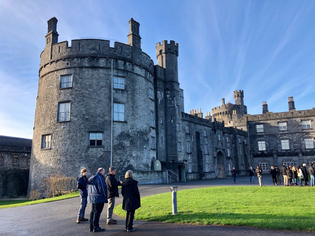 Eine Gruppe von Menschen steht vor dem Kilkenny Castle, das von grünem Gras und Pflanzen umgeben ist und an einem teilweise bewölkten Himmel liegt.