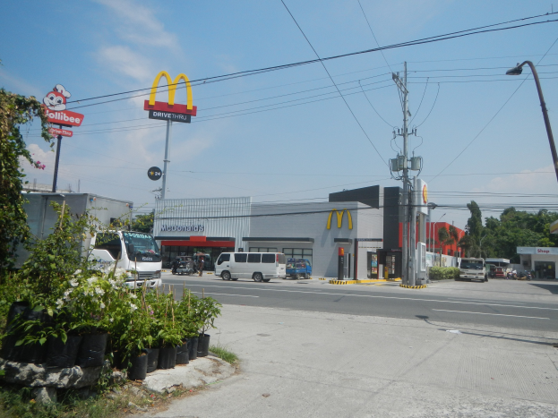 Ein McDonald's-Restaurant an einer Straßenecke mit vorbeifahrenden Fahrzeugen, umgeben von Gebäuden, Straßenelementen, Grünflächen und einem bewölkten Himmel.