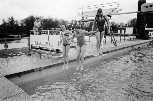 Schwarzes und weißes Foto von drei Frauen, die in einen Pool springen, umgeben von Bäumen und Gebäuden unter einem sichtbaren Himmel.