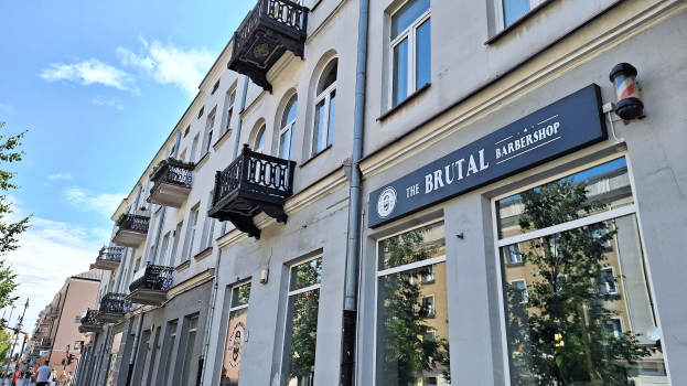 Eine Straßenansicht eines belebten Friseurladens in Berlin, Deutschland, mit umliegenden Gebäuden, die Fenster, Geländer, Balkone und Schilder tragen, Bäumen, Laternenpfählen und Menschen, die auf der Straße unter einem bewölkten Himmel gehen.