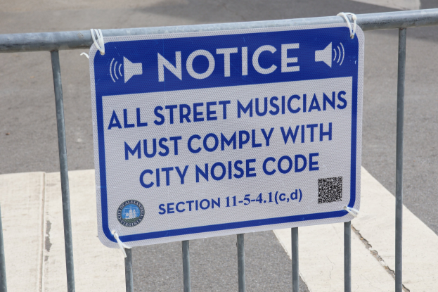 Anschlagtafel an einem Zaun mit einem Schild, auf dem "Anschlag: Alle Straßenmusiker müssen der städtischen Lärmvorschrift Folge leisten" steht, und einer Straße im Hintergrund.