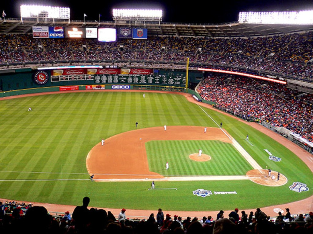 Baseball stadium with players on the field, hoardings, lights, flags, and an audience in the background.