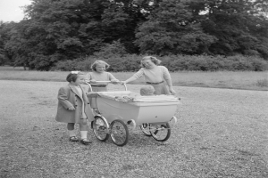 Drei junge Mädchen schieben einen Kinderwagen mit einem Baby darin, umgeben von Bäumen unter einem klaren Himmel, dargestellt in Schwarz-Weiß.