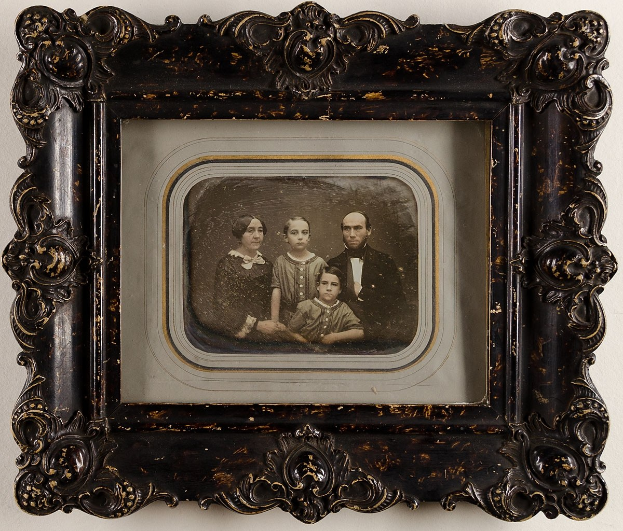 Altes Familienfoto in einem prunkvoll verzierten Rahmen auf einer weißen Oberfläche, mit der Familie, die in die Kamera lächelt.