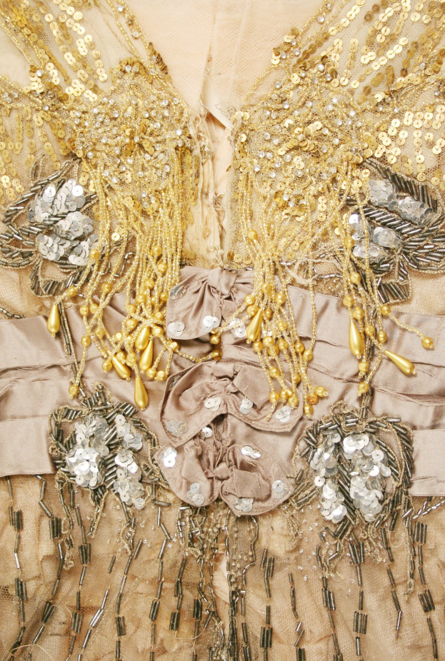 Ein Close-up eines hellen Kleides mit goldenen Pailletten und Perlen, das im Licht funkelt.