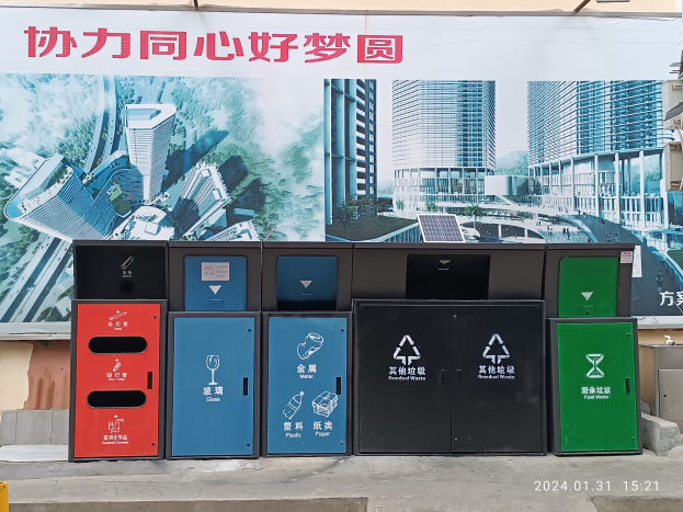 Eine Reihe bunter Mülltonnen, die entlang einer Straße aufgereiht sind, mit einem Banner dahinter, einem Objekt auf dem Boden rechts daneben und einem Wasserzeichen, das auf Hongkongs Müllentsorgungssystem hinweist.