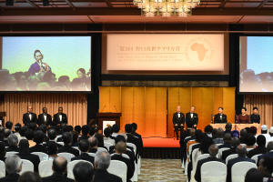 Eine Gruppe von Menschen sitzt in einem Raum mit einem Podium, Mikrofonen und einem großen Bildschirm, der Text und Bilder anzeigt, mit einer von einem Kronleuchter beleuchteten Decke darüber.