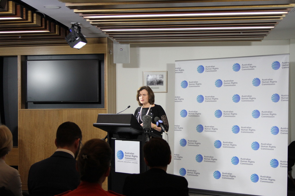 Eine Frau steht auf einer Bühne und spricht in ein Mikrofon, vor ihr sitzt ein Publikum, an der Wand hängt ein Banner, auf dem Holztisch steht ein Fernseher und an der Decke sind Lampen.