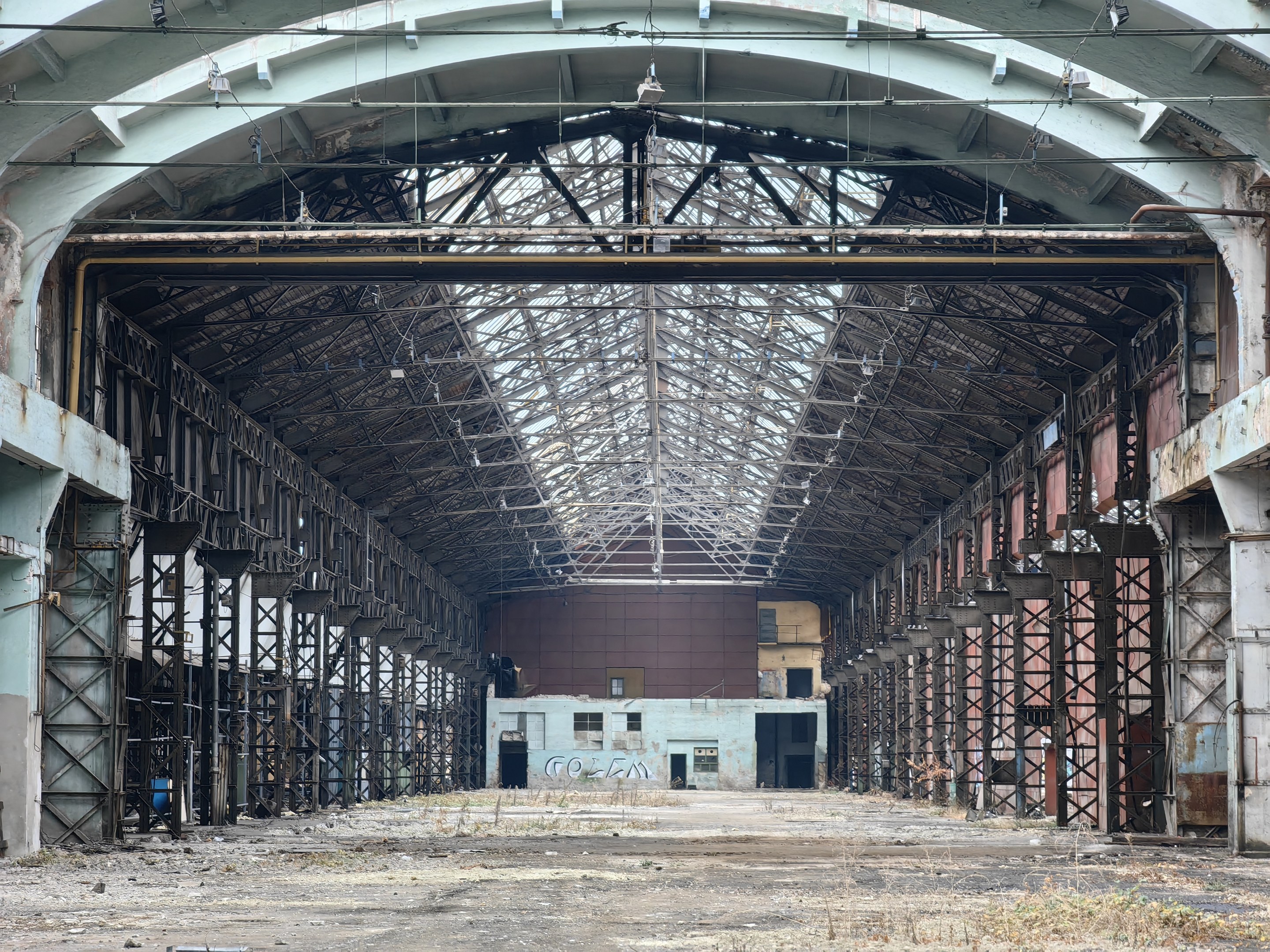 Old abandoned building with numerous windows, rusted metal poles, a deteriorating roof with ceiling lights, and a text-covered wall, showing signs of decay and disrepair.