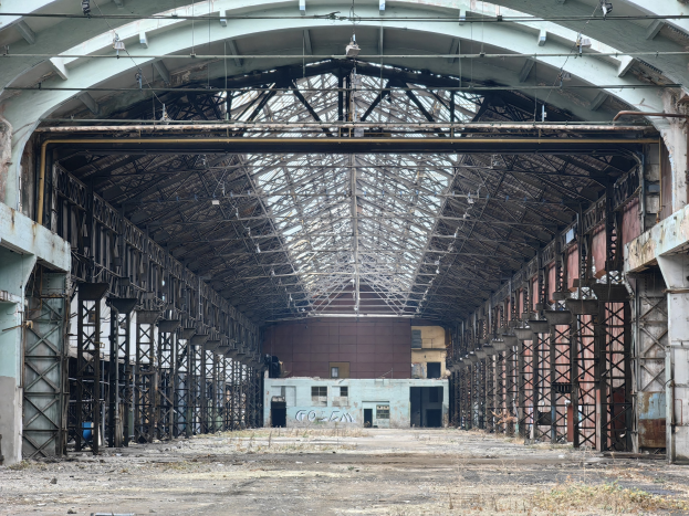 Old abandoned building with numerous windows, rusted metal poles, a deteriorating roof with ceiling lights, and a text-covered wall, showing signs of decay and disrepair.
