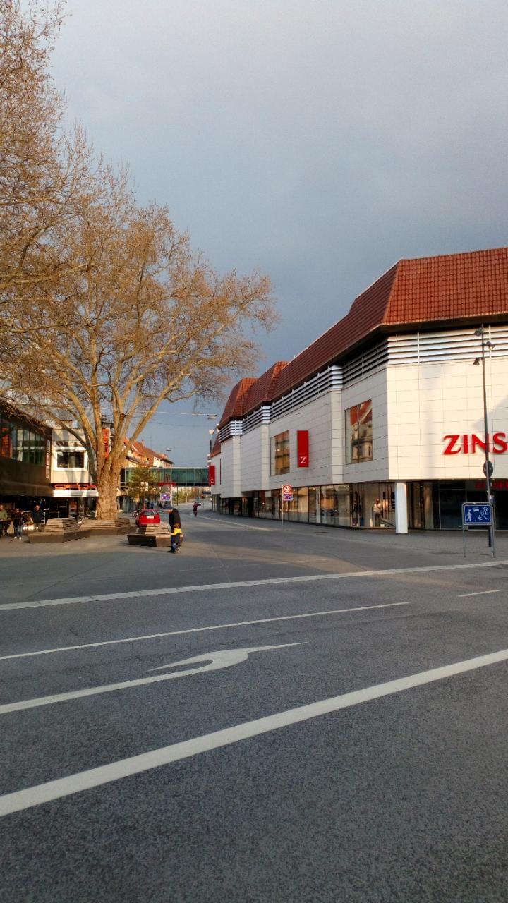 Eine Straße in Berlin, Deutschland, mit einem roten und weißen Zins-Einkaufszentrum, umgeben von Bäumen, Pfählen und Menschen, mit einem sichtbaren Himmel im Hintergrund.