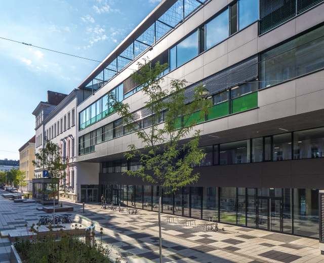 Großes modernes Gebäude mit vielen Fenstern, umgeben von Bäumen, Pflanzen, Fahrzeugen und Menschen unter einem bewölkten Himmel, identifiziert als das neue Hauptquartier des Deutschen Instituts für Technologie in Berlin.