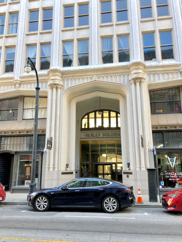 Ein Tesla Model S vor einem großen Gebäude mit Glasfenstern und -türen geparkt, ein Schild, ein Straßenpfosten, ein Verkehrszeichen und Autos auf der Straße.