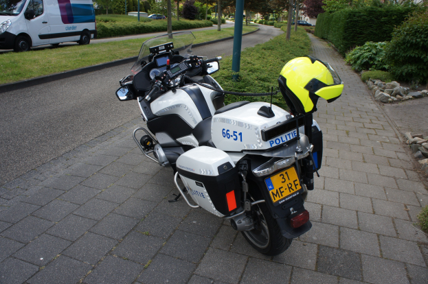 Ein Polizeimotorrad am Straßenrand geparkt mit einem Helm darauf, umgeben von Bäumen, Pfählen, Gebäuden, Pflanzen, Gras, Steinen und anderen Fahrzeugen.