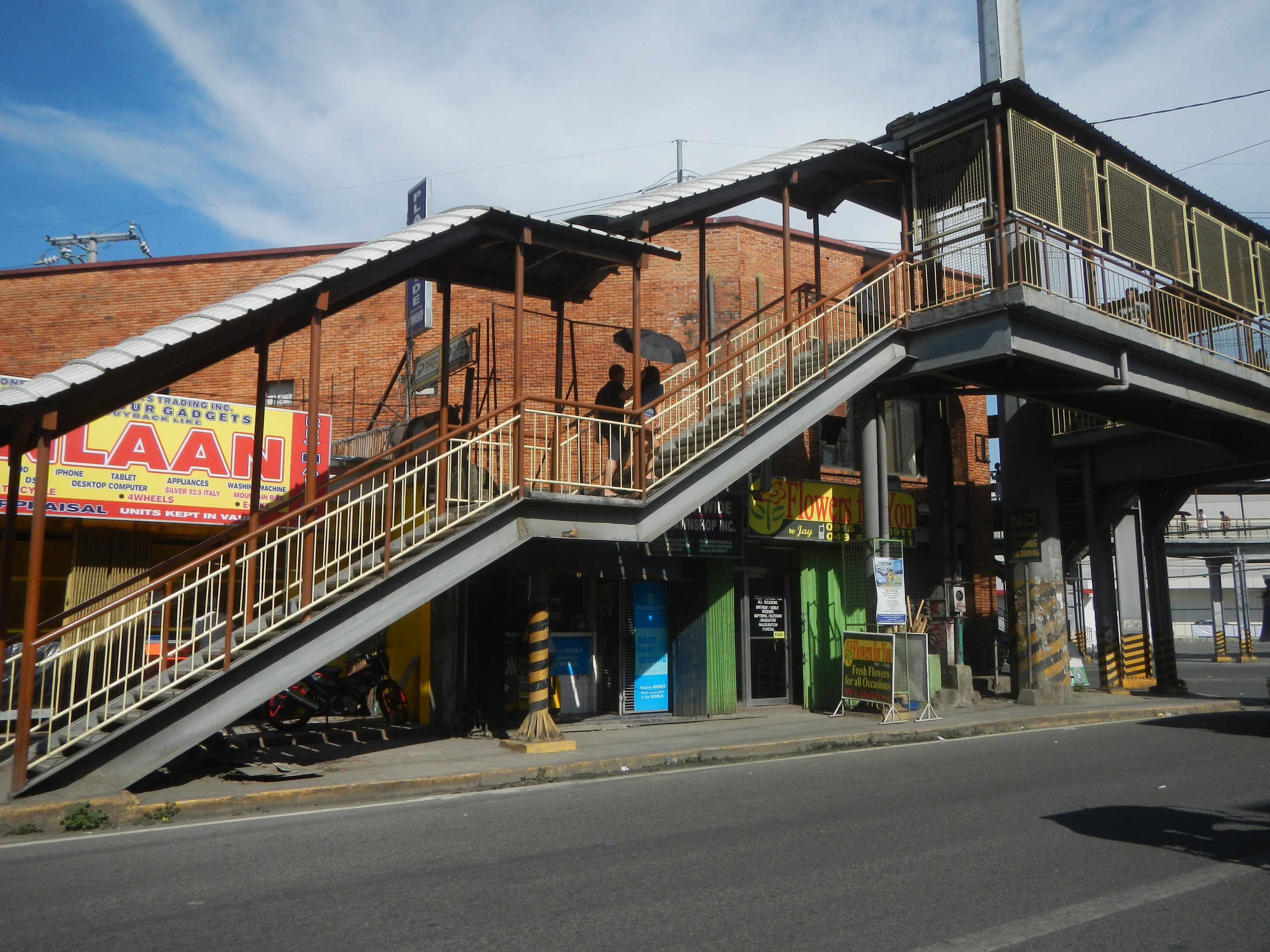 Ein Bahnhofsgebäude mit einem Zug auf den Gleisen, eine Brücke mit Geländern und Treppen, Gebäude mit Namensschildern, geparkte Motorräder, Strommasten mit Kabeln und ein bewölkter Himmel im Hintergrund.