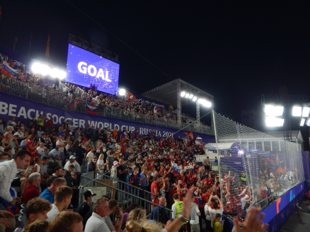 Große Menschenmenge versammelt sich nachts vor einem Beachsoccer-Tor mit Netz auf der rechten Seite und einem beleuchteten Bildschirm im Hintergrund.