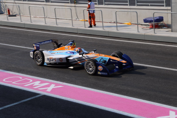 Ein Mann fährt ein Formelauto auf einer Rennstrecke, mit einer anderen Person auf dem Fahrersitz, die einen Helm trägt, und einer Person im Hintergrund auf dem Bürgersteig.