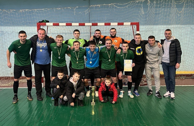 Gruppe von Männern in Sportbekleidung auf einem Futsal-Platz stehend, die Medaillen tragen und einen Pokal halten, mit einem Tor und einer Wand im Hintergrund.