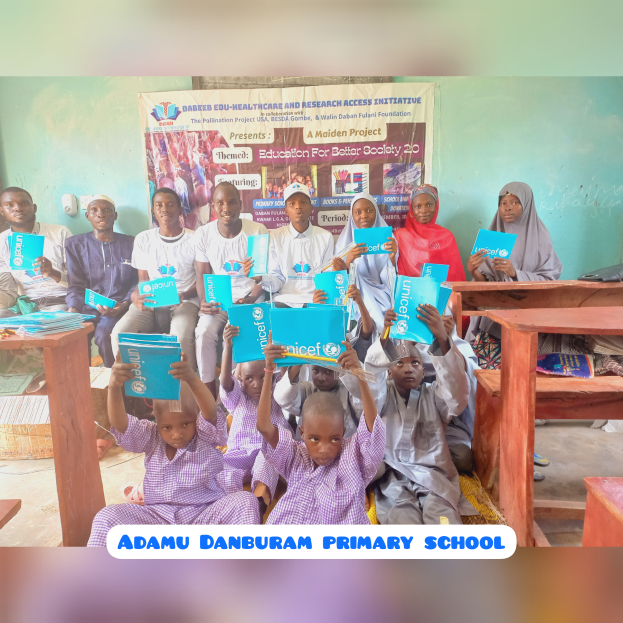 Eine Gruppe von Kindern sitzt auf Bänken in einem Klassenzimmer, hält Bücher in den Händen, mit Tischen davor, die Bücher und andere Gegenstände enthalten, und einem Banner an der Wand mit der Aufschrift "Adamu Danburam Grundschule".