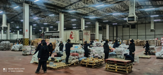 Eine Gruppe von Menschen steht in einem großen Lagerhaus voller Lebensmittelvorräte, Holzpaletten, Container, und strukturelle Elemente, was auf ein Lebensmittelverteilungszentrum in Dubai hinweist.