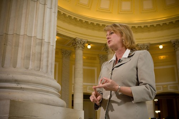 Frau in einem Mantel vor einer beleuchteten Säule in einem Gebäude stehend, die darauf hinweist, dass sie Politikerin ist.