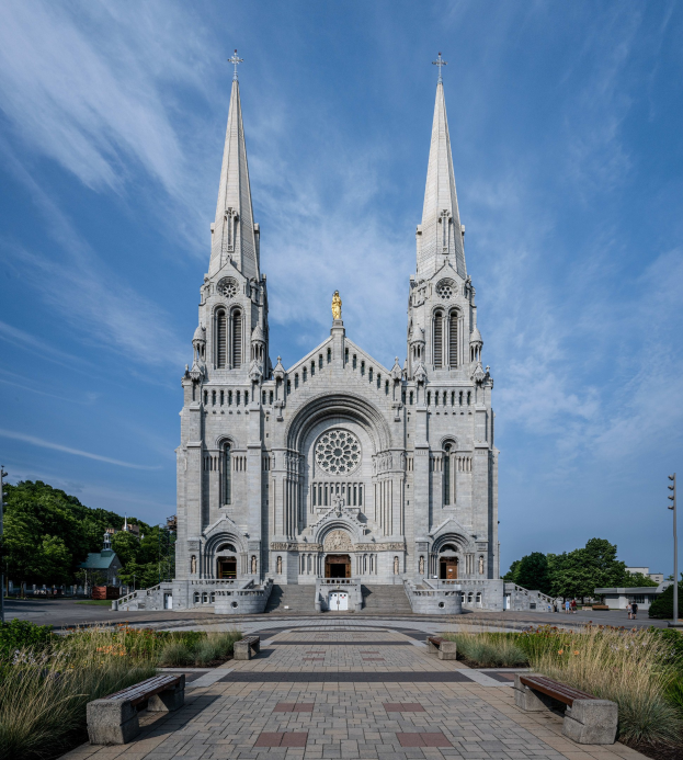 Majestätische St. Patrick's Cathedral in Dublin, Irland mit zwei Türmen, Statuen, Fenstern, Türen, Stufen und Geländern, vor denen Bänke stehen, flankiert von Pflanzen und Gras sowie Bäumen, Laternenmasten und einem bewölkten Himmel im Hintergrund.