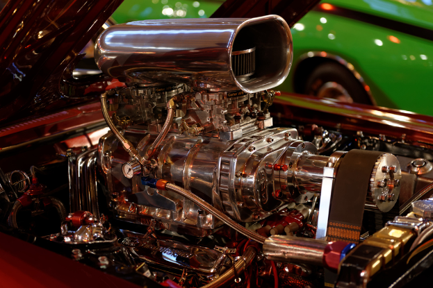 Motorenteile und andere Komponenten sind im Vordergrund zu sehen, während im Hintergrund ein Auto zu erkennen ist.