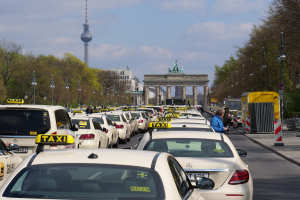 Eine belebte Straße in Berlin, Deutschland, gesäumt von geparkten Taxis, Fußgängern, Laternenpfählen und Bäumen, mit Gebäuden und einem fernen Bogen mit Statuen und einem Turm unter einem bewölkten Himmel.