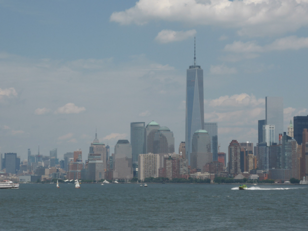 Stadtpanorama-Ansicht vom Wasser aus mit Booten im Vordergrund und dem One World Trade Center im Hintergrund, umgeben von Bäumen und Wolken.