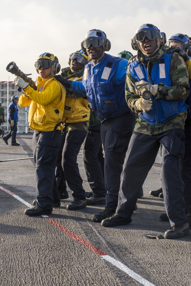 Gruppe von Männern in Helmen und Schutzbrillen, die auf einem Flugzeugträger stehen, einige halten Waffen, mit Gebäuden und einem klaren blauen Himmel im Hintergrund.