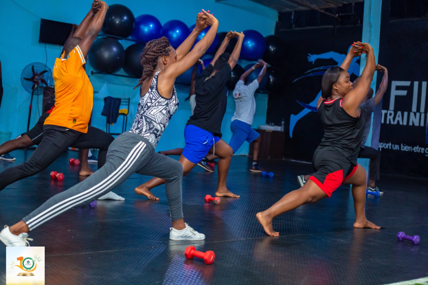Gruppe von Menschen, die Yoga in einem Fitnessstudio mit Hanteln auf dem Boden machen, mit einem Banner im Hintergrund, auf dem 'Fitness-Training in Ghana' steht.