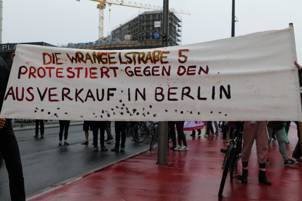 Eine Gruppe von Menschen hält ein Transparent mit der Aufschrift "Die Wrangellstraße 5 Protestiert Gegen den Ausverkauf in Berlin" auf einer Straße, mit Fahrrädern, Pfählen, Gebäuden und einem Kran im Hintergrund.