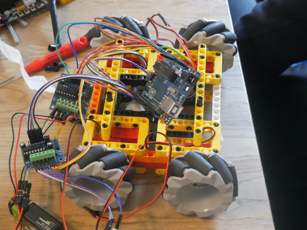 Ein Holztisch mit elektronischen Bauteilen, darunter ein Arduino Uno R3 Roboter, der über Kabel verbunden ist, neben einem Menschenbein.
