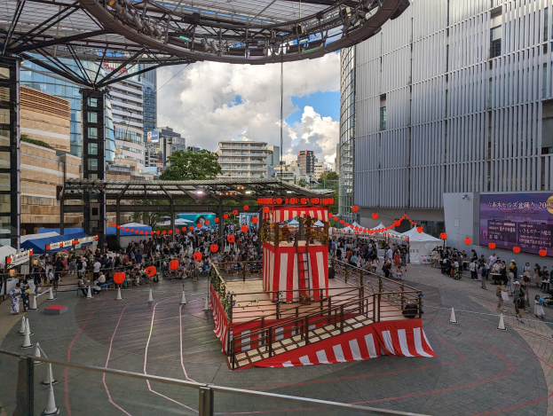 Große Menschenmenge versammelt sich auf einer Bühne in einer städtischen Umgebung zur Eröffnung des Tokyo Circus Festivals, mit Performern und Equipment im Bild.