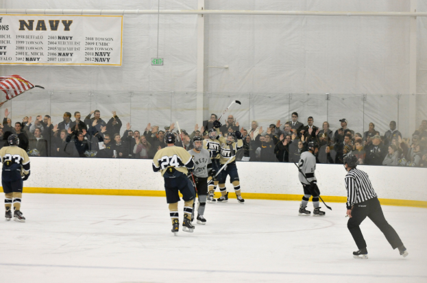 Eishockeyspieler in Helmen und Uniformen konkurrieren auf einem Eisstadion mit Zuschauern in den Rängen und einer Bannerwand im Hintergrund.