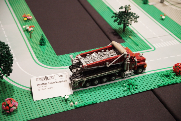 Ein Modell-Lkw aus Bausteinen, das zwischen Bäumen, Blumen und anderen Gegenständen ausgestellt ist, mit einer Texttafel im Hintergrund.