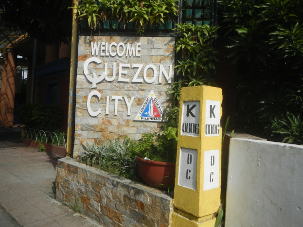 Willkommensschild für Quezon City, Philippinen, mit Text an der Wand, Topfpflanzen, einem Metallzaun, Bäumen, einem Gebäude mit Fenstern und einem Dach.