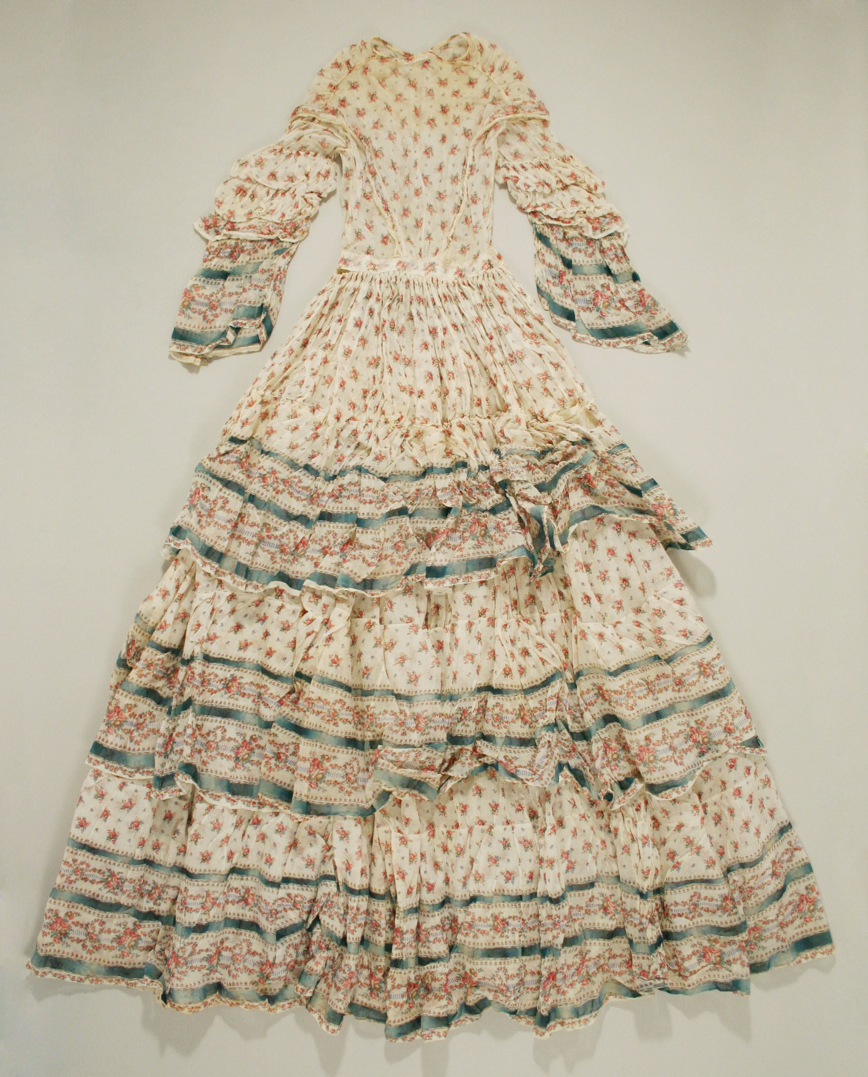 White floral-patterned dress with a fitted bodice, full floor-length skirt, and cascading ruffles hanging on a wall.
